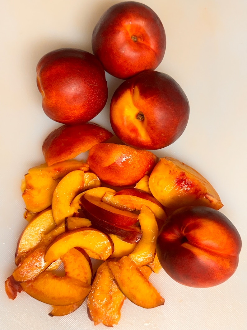 May include: A white cutting board with four whole nectarines and several slices of nectarine. The nectarines are red and orange with a yellow center. The slices are yellow and orange.