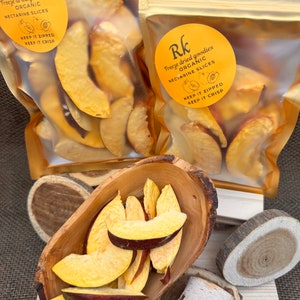 May include: Two bags of freeze-dried organic nectarine slices. The bags are gold with a yellow label that says "Freeze dried goodies, Organic, Nectarine Slices, Keep it zipped, Keep it crisp". A wooden bowl filled with the nectarine slices is in the foreground.