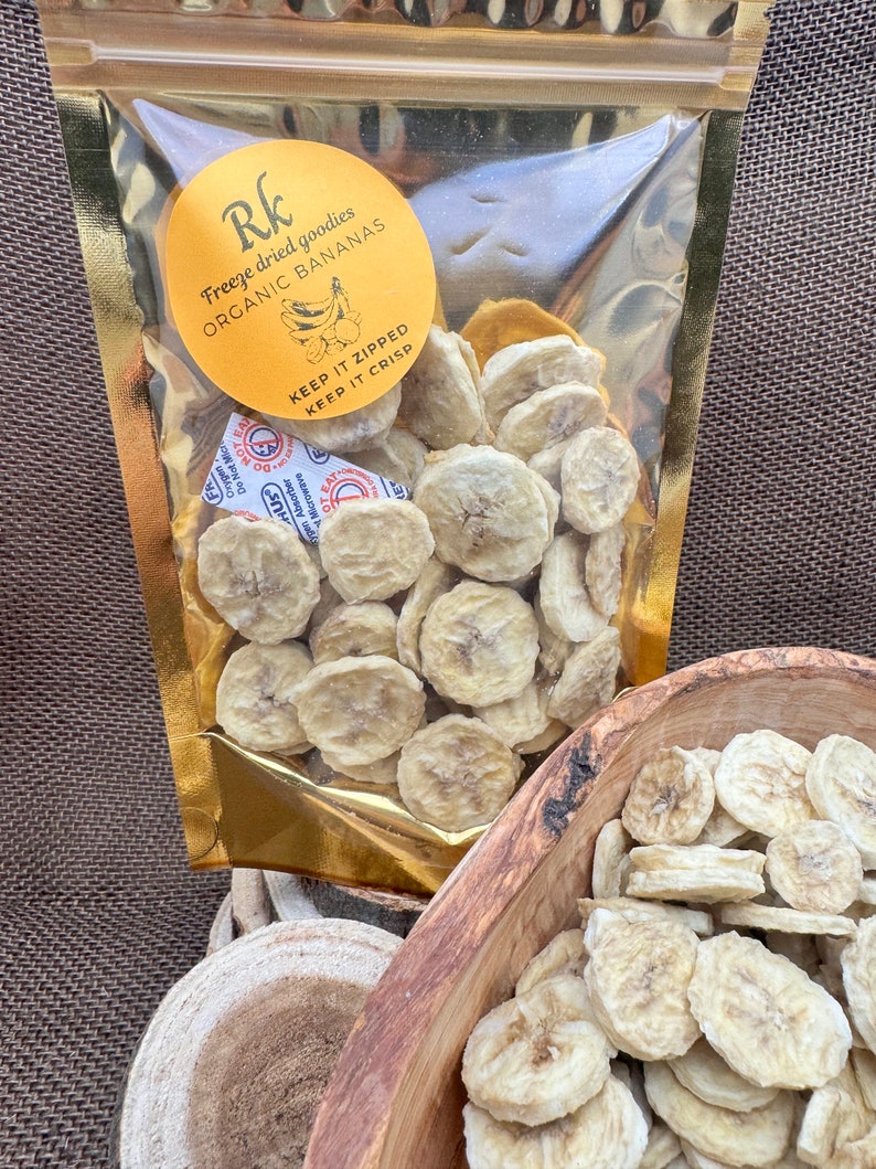 May include: A gold resealable bag filled with freeze-dried organic banana slices. The bag has a yellow label with the text "Rk Freeze dried goodies ORGANIC BANANAS KEEP IT ZIPPED KEEP IT CRISP".