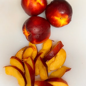 May include: Three whole red and yellow nectarines and several slices of nectarine on a white cutting board.