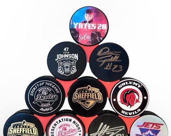 May include: A stack of black and white hockey pucks with various logos and designs. The top puck features a player in a red and white jersey with the text "Yates 28" and "Sola". Other pucks feature logos for "Junior Sheffield", "Spirit of the Game", "Sulent Devils", and "Jets".