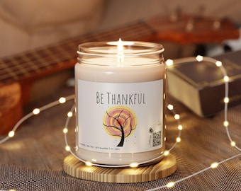 May include: A lit candle in a clear glass jar with a label that reads "BE THANKFUL" and a tree graphic. The candle sits on a round wooden coaster, with string lights and a guitar in the background.