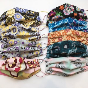 Op de afbeelding: Een set van acht kleurrijke stoffen mondmaskers met verschillende patronen, waaronder bloemen, geometrische en schedelontwerpen. De maskers zijn geplooid en hebben elastische oorlussen.