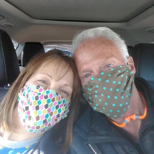 Op de afbeelding: Een stel dat gezichtsmaskers draagt in een auto. De vrouw draagt een roze, groene en blauwe gebloemde masker. De man draagt een bruin masker met turquoise stippen.