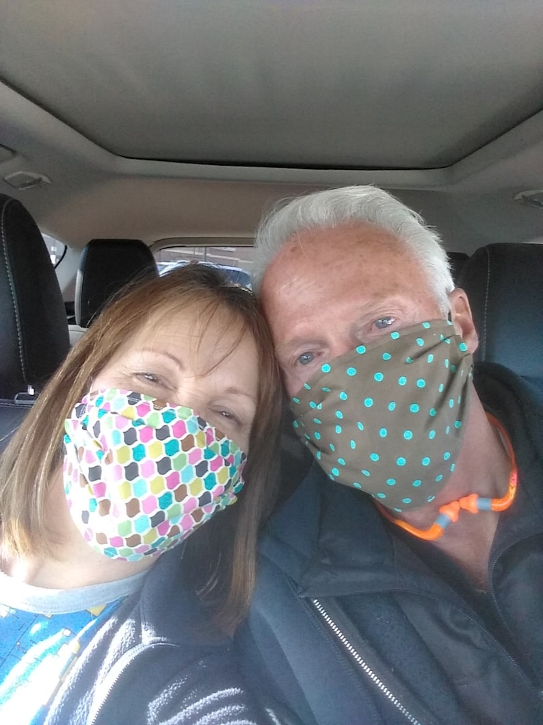 Op de afbeelding: Een stel dat gezichtsmaskers draagt in een auto. De vrouw draagt een roze, groene en blauwe gebloemde masker. De man draagt een bruin masker met turquoise stippen.