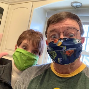 Op de afbeelding: Twee mensen dragen gezichtsmaskers. De persoon links draagt ​​een groen masker met witte stippen. De persoon rechts draagt ​​een blauw masker met een kleurrijk patroon van bomen en een hut.
