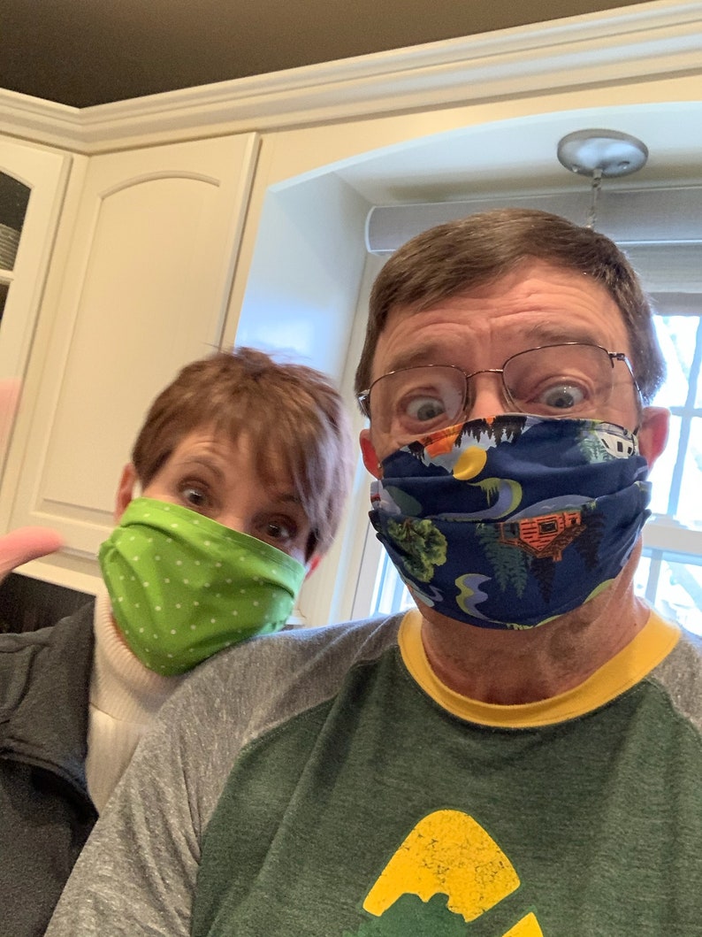 Op de afbeelding: Twee mensen dragen gezichtsmaskers. De persoon links draagt ​​een groen masker met witte stippen. De persoon rechts draagt ​​een blauw masker met een kleurrijk patroon van bomen en een hut.
