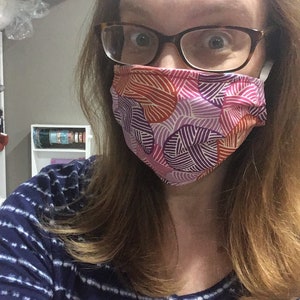 Op de afbeelding: Een vrouw draagt ​​een mondkapje met een kleurrijk geometrisch patroon. Het masker is gemaakt van stof en heeft een roze, paarse en oranje print. De vrouw draagt ​​ook een bril.