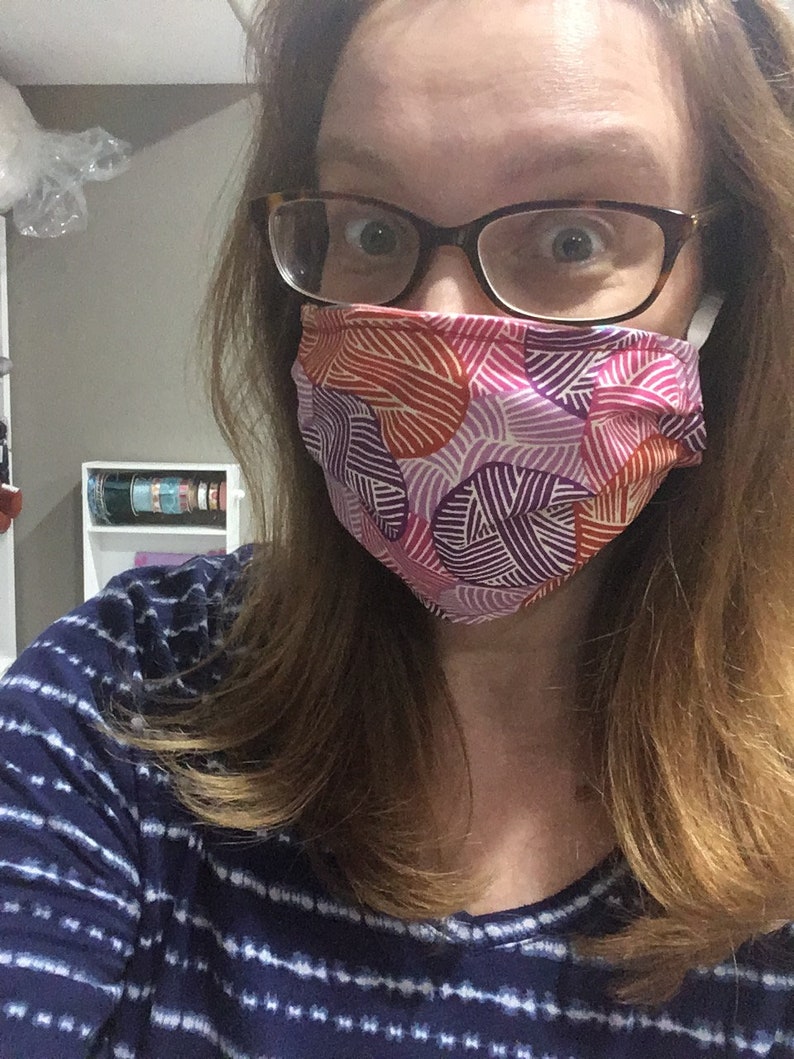 Op de afbeelding: Een vrouw draagt ​​een mondkapje met een kleurrijk geometrisch patroon. Het masker is gemaakt van stof en heeft een roze, paarse en oranje print. De vrouw draagt ​​ook een bril.
