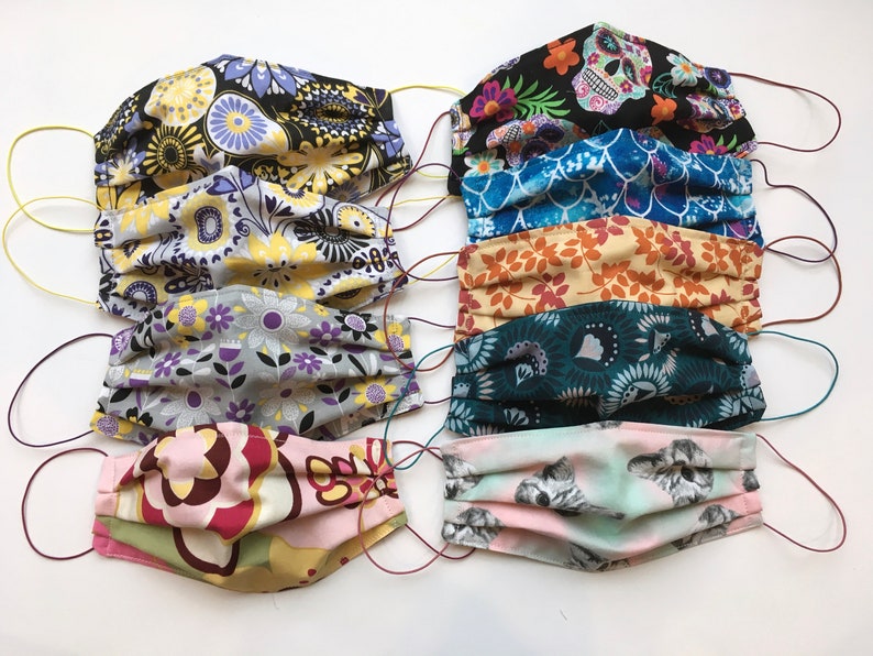Op de afbeelding: Een set van acht kleurrijke stoffen mondmaskers met verschillende patronen, waaronder bloemen, geometrische en schedelontwerpen. De maskers zijn geplooid en hebben elastische oorlussen.