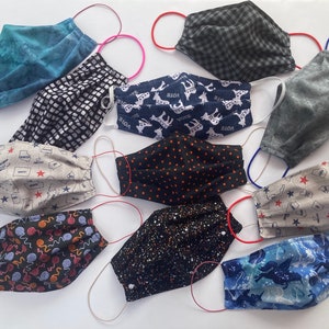 Op de afbeelding: Een verzameling van 10 stoffen mondmaskers met verschillende patronen en kleuren, waaronder blauw, zwart, wit, grijs en rood. De maskers zijn gemaakt van katoen en hebben elastische oorlussen. Sommige maskers hebben bedrukte ontwerpen, zoals sterren, stippen en dieren.