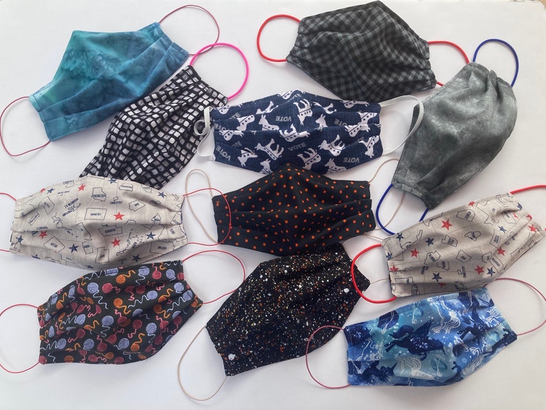Op de afbeelding: Een verzameling van 10 stoffen mondmaskers met verschillende patronen en kleuren, waaronder blauw, zwart, wit, grijs en rood. De maskers zijn gemaakt van katoen en hebben elastische oorlussen. Sommige maskers hebben bedrukte ontwerpen, zoals sterren, stippen en dieren.