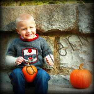 May include: A young boy wearing a grey sweater with a red and white design sits on a stone wall. He is holding two small pumpkins in his hands. The pumpkins are orange and round.