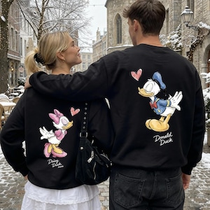 May include: Black sweatshirts featuring Disney's Daisy Duck and Donald Duck characters. Daisy Duck is depicted in pink and white, while Donald Duck is in blue and white. Each design includes a pink heart and character name.