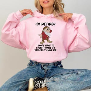 May include: A light pink hoodie with the text "I'M RETIRED" and a cartoon image of a dwarf. Below the image is the text "I DON'T HAVE TO I DON'T WANT TO YOU CAN'T MAKE ME". The hoodie is made of a soft material.