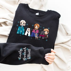 Puede incluir: Sudadera negra con un gr&aacute;fico de los personajes de la pel&iacute;cula Frozen, incluyendo Olaf, Elsa, Anna y Kristoff. El texto "MAMA" est&aacute; escrito en una fuente estilizada con copos de nieve. La manga de la sudadera tiene los nombres "Emily, Hannah, Elliott" escritos en blanco con copos de nieve.