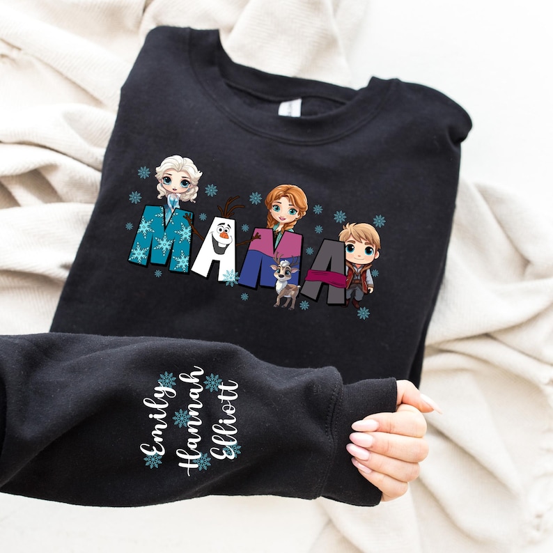 Puede incluir: Sudadera negra con un gr&aacute;fico de los personajes de la pel&iacute;cula Frozen, incluyendo Olaf, Elsa, Anna y Kristoff. El texto "MAMA" est&aacute; escrito en una fuente estilizada con copos de nieve. La manga de la sudadera tiene los nombres "Emily, Hannah, Elliott" escritos en blanco con copos de nieve.