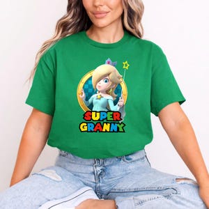 May include: A bright green short-sleeved t-shirt featuring a graphic of the character Rosalina from the Super Mario series inside a gold ring. Below the character, the text SUPER GRANNY is printed in a bold, multi-coloured, blocky font style.