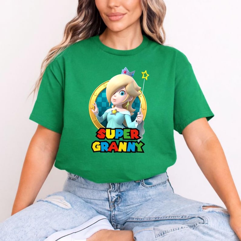May include: A bright green short-sleeved t-shirt featuring a graphic of the character Rosalina from the Super Mario series inside a gold ring. Below the character, the text SUPER GRANNY is printed in a bold, multi-coloured, blocky font style.