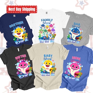 May include: Set of six t-shirts featuring Baby Shark characters. The shirts are in different colors and feature the text "Brother Shark", "Family Shark", "Dad Shark", "Sister Shark", "Baby Shark", and "Mom Shark". Each shirt has a different Baby Shark character and the text "doo doo doo doo doo doo".