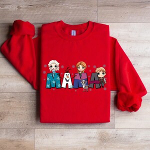 Puede incluir: Sudadera roja con un gr&aacute;fico de los personajes de Frozen, incluyendo Elsa, Anna, Olaf y Kristoff, deletreando la palabra "MAMA" con copos de nieve.