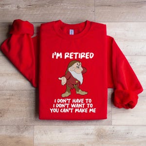 May include: A red sweatshirt with the text "I'M RETIRED" in white, along with a cartoon image of a grumpy dwarf. Below the image, the text reads "I DON'T HAVE TO I DON'T WANT TO YOU CAN'T MAKE ME".