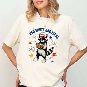 May include: A white t-shirt with a graphic of a raccoon wearing a red, white, and blue "Merica" hat and holding a hot dog. The graphic is surrounded by stars and flowers. The text "Red White and Feral" is printed in a circle around the raccoon.