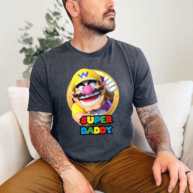 May include: A dark heather grey short-sleeved t-shirt featuring a graphic of the character Wario inside a gold circle. Below the character, the text reads SUPER DADDY in bold, colourful block letters. The shirt has a crew neckline and a standard fit.