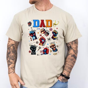 May include: Beige t-shirt with the word "DAD" in blue, orange, and red. The shirt features illustrations of superhero fists with words like "devoted," "fearless," and "strong."