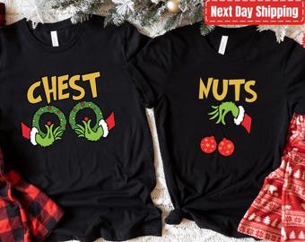 May include: Two black t-shirts with Christmas-themed graphics. One shirt says "CHEST" with green wreath designs and Grinch-like hands. The other says "NUTS" with Grinch hands and red ornament graphics. Festive holiday apparel.