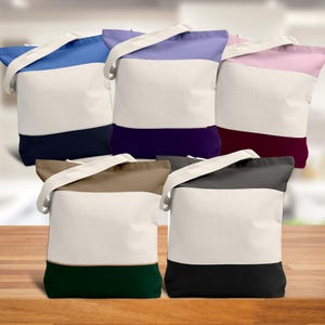 May include: Five canvas tote bags in various colors: blue, purple, pink, brown, and gray. Each bag has a white center panel, a colored top and bottom, and a cream-colored strap. The bags are displayed on a wooden surface.