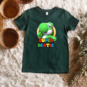 May include: A dark green short-sleeved t-shirt featuring a graphic of the green character Yoshi from the Super Mario series inside a gold circle. Below the character, the text SUPER BESTIE is printed in a multi-coloured, blocky, pixelated font.