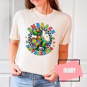 May include: A white t-shirt with a colorful graphic of a dinosaur wearing sunglasses and holding a puzzle piece heart. The graphic is surrounded by a circle with the text "Embrace the Differences". The shirt is available in ivory.