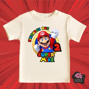 May include: A cream-coloured short-sleeve t-shirt featuring a graphic of Mario inside a gold ring. The text reads "BIRTHDAY BOY", "SUPER MIKE", and the number "2" in red, blue, yellow, and green colours. The design is centred on the front of the shirt.