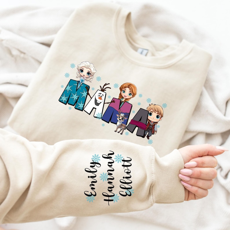 Puede incluir: Una sudadera beige con un gr&aacute;fico de los personajes de Frozen deletreando "MAMA" con copos de nieve. La manga tiene los nombres "Emily, Hannah, Elliott" con copos de nieve.