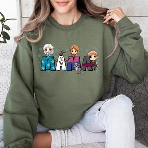 Puede incluir: Sudadera verde con un gr&aacute;fico de los personajes de Frozen, incluyendo Elsa, Anna, Olaf, Kristoff y Sven. El gr&aacute;fico dice "MAMA" con cada letra representando un personaje diferente.