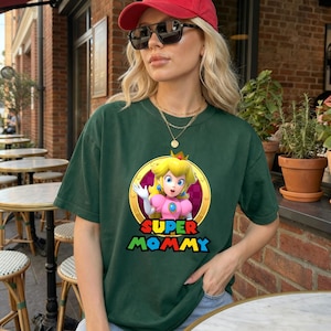 May include: A dark green short-sleeved t-shirt featuring a graphic of Princess Peach inside a gold circular frame. Below the character, the text reads SUPER MOMMY in bold, colourful, block-style lettering. The shirt has a crew neckline.