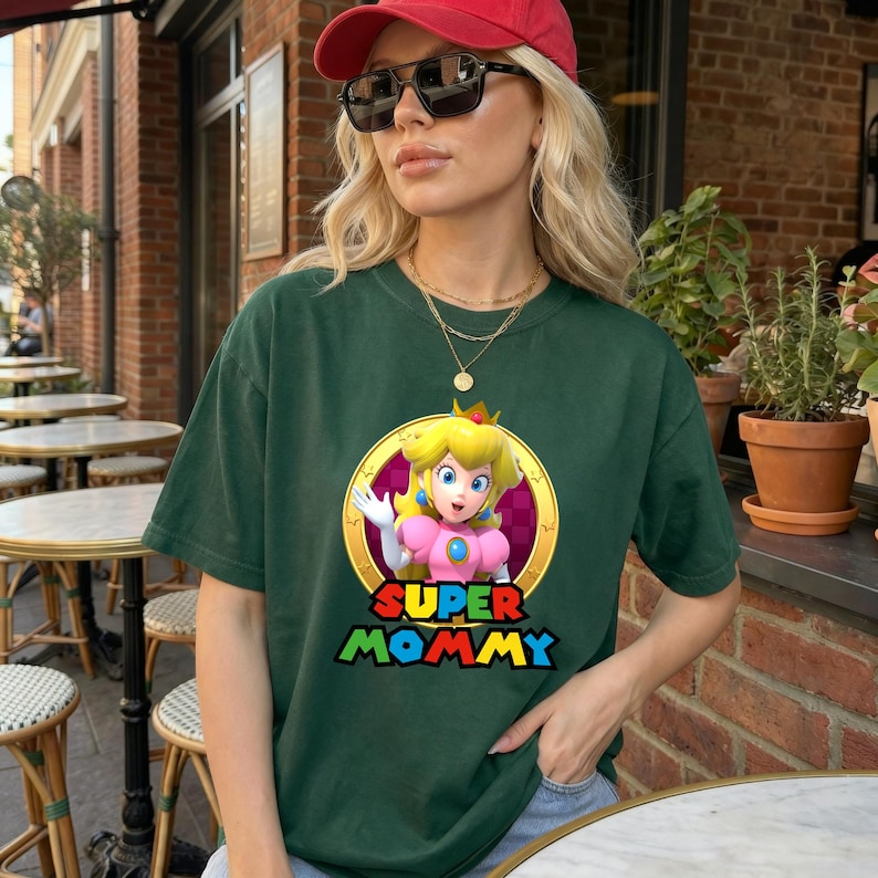 May include: A dark green short-sleeved t-shirt featuring a graphic of Princess Peach inside a gold circular frame. Below the character, the text reads SUPER MOMMY in bold, colourful, block-style lettering. The shirt has a crew neckline.