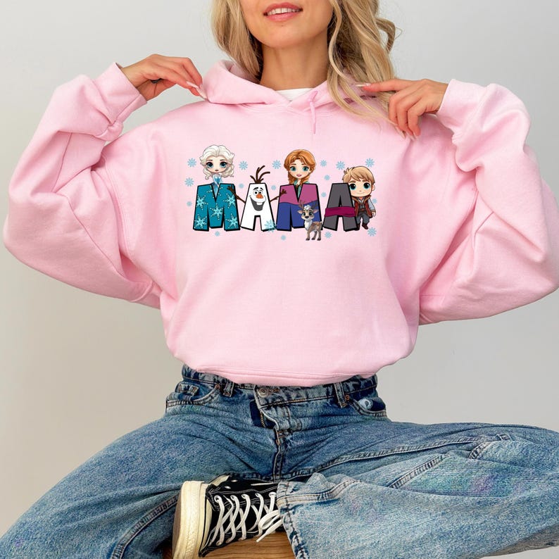 Puede incluir: Sudadera rosa con capucha con un estampado gr&aacute;fico de personajes de la pel&iacute;cula Frozen. El gr&aacute;fico incluye la palabra "MAMA" con los personajes de pie sobre las letras.