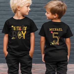 May include: Two black t-shirts with a construction theme. One shirt says "Birthday Boy" with yellow and black construction vehicles and tools. The other shirt has a large yellow number 5 with black stripes and construction vehicles and tools. The shirt also has the name "Michael" printed on it.