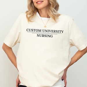 May include: Cream-colored t-shirt with the text "CUSTOM UNIVERSITY NURSING" in black, centered on the front. The text is underlined. The shirt has short sleeves and a crew neck.