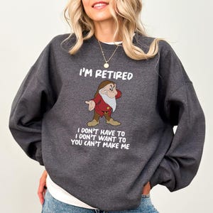 May include: Dark gray sweatshirt with the text "I'M RETIRED" and a cartoon image of a grumpy dwarf. Below the image, the text reads "I DON'T HAVE TO I DON'T WANT TO YOU CAN'T MAKE ME". The sweatshirt has a crew neck.