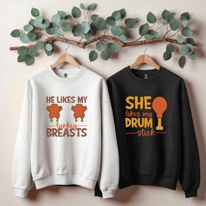 May include: Two sweatshirts on wooden hangers. One is white with the text "HE LIKES MY turkey BREASTS" and a turkey graphic. The other is black with the text "SHE likes my DRUM stick" and a drumstick graphic. A Thanksgiving theme.