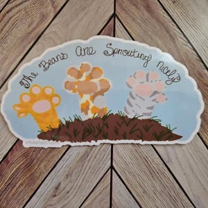May include: A sticker with a light blue background showing three cat paws emerging from brown soil. The paws are orange, orange and white, and grey and white. The text "The Beans are Sprouting nicely" is written above the paws.