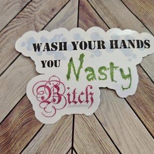 Puede incluir: Una pegatina blanca con el texto "WASH YOUR HANDS YOU Nasty Bitch" en letras negras, verdes y rojas. La pegatina tiene forma de nube con burbujas en la parte superior. La pegatina está sobre una superficie de madera.
