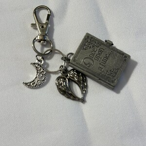 May include: A silver-toned keychain with a lobster clasp, featuring charms of a crescent moon, angel wings, and a small book. The book charm is engraved with the words "Once upon a time..."