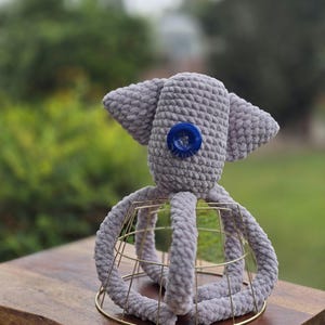 Op de afbeelding: Een handgemaakt grijs gehaakt octopus speelgoed met een blauwe knoop als oog. De octopus staat op een gouden draadframe. Het pluche speelgoed heeft grote, slappe oren en dikke, afgeronde tentakels.