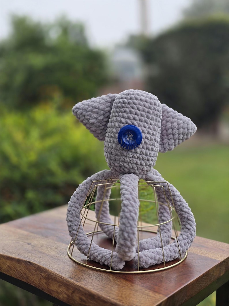 Op de afbeelding: Een handgemaakt grijs gehaakt octopus speelgoed met een blauwe knoop als oog. De octopus staat op een gouden draadframe. Het pluche speelgoed heeft grote, slappe oren en dikke, afgeronde tentakels.