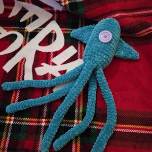 Op de afbeelding: Een turquoise gehaakte octopus knuffel met een lavendelkleurig knoopoog. De knuffel heeft acht lange tentakels en ligt op een rood-zwarte geruite deken met witte tekst.