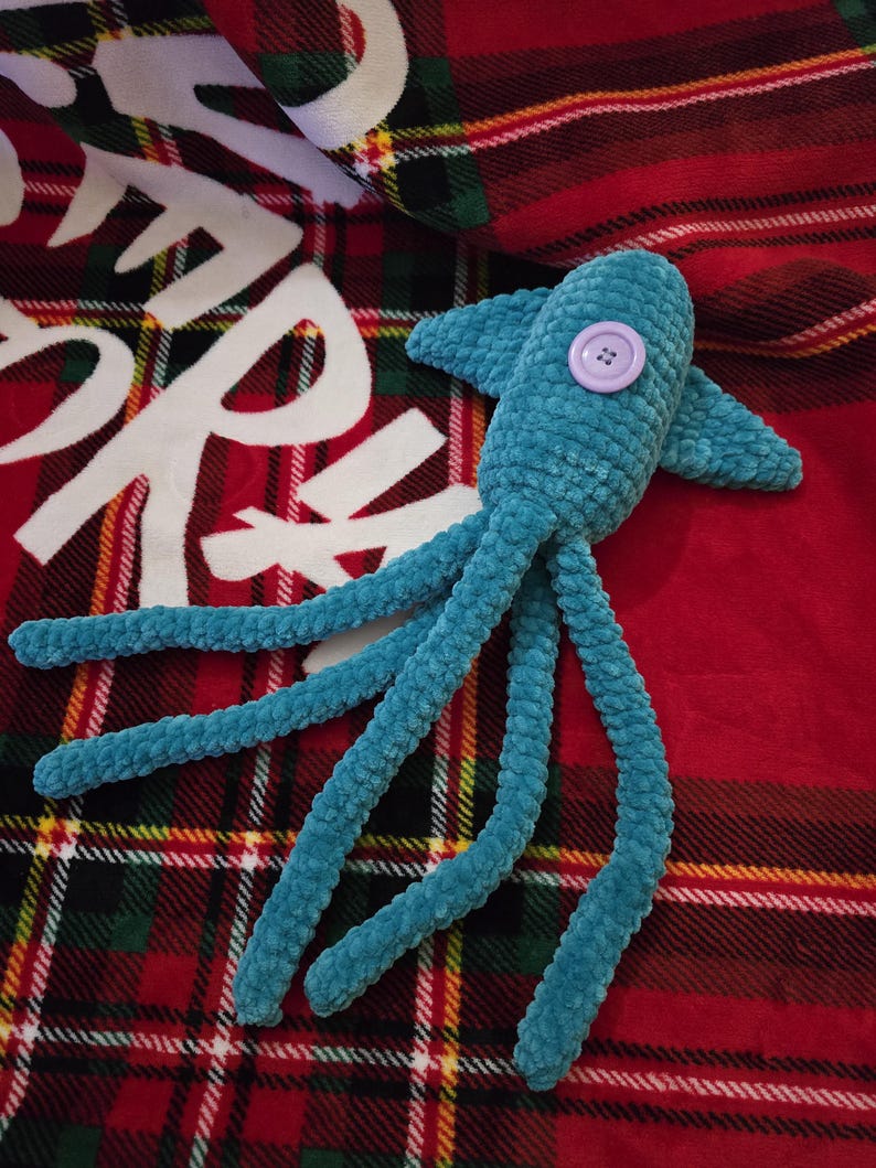 Op de afbeelding: Een turquoise gehaakte octopus knuffel met een lavendelkleurig knoopoog. De knuffel heeft acht lange tentakels en ligt op een rood-zwarte geruite deken met witte tekst.